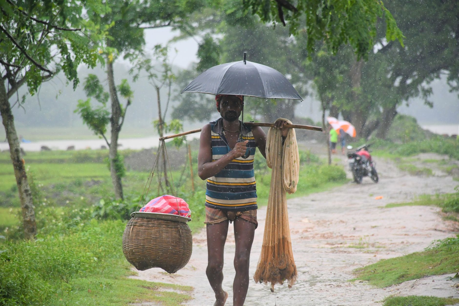 A fisherman walks outdoors in the rain with an umbrella in Pirganj, Bangladesh, carrying traditional fishing gear.